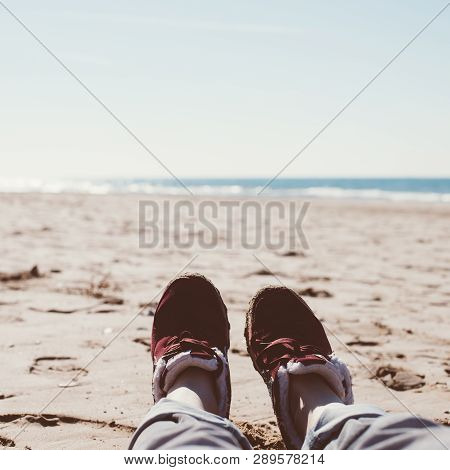 Feet Of Young Woman In Sport Footwear And Jeans Relaxing After Hiking On Sand Beach. Square Pov. Adv