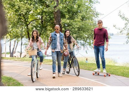 Handsome African American Man Walking Near Pretty Girls Riding Bicycles And Cheerful Friend Longboar