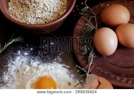 Eggs, Flour And Ovine Cereal Lie On A Clay Tray. Dark Background. Ingredients For Cooking Homemade, 