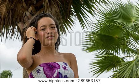 Young Girl With Floral Sundress Smiling With Flower In Hair Standing Under Palm Tree