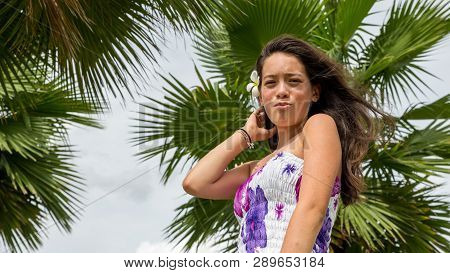 Young Girl With Floral Sundress Smiling With Flower In Hair Standing Under Palm Tree