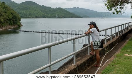 Asian Woman Leaning On Railing Looking At Resevoir With Mountains In The Back Ground