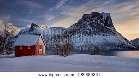 Red Cabin In Snow At Steinfjorden With Frozen Mountains In Background, Senja, Norway