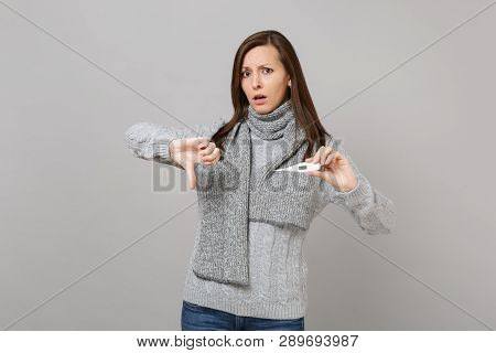 Puzzled Young Woman In Gray Sweater Scarf Showing Thumb Down, Hold Thermometer Isolated On Grey Wall