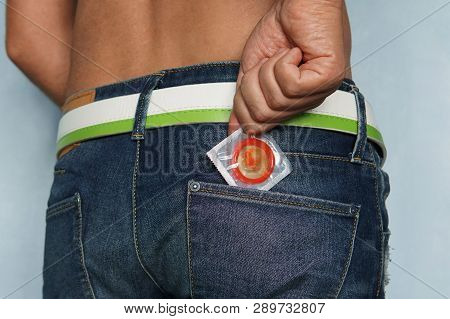 Back Shot Of Young Male On Blue Background Taking The Latex Condom Out Of Jeans Pocket. Half-dressed