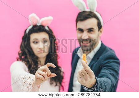 Young Beautiful Couple Standing On A Pink Background. During This, A Man Holds A Small Carrot In His