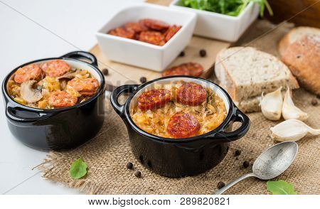 Slovak Christmas National Cabbage Soup In Two Small Black Pots With Sausage On The Jute Background.