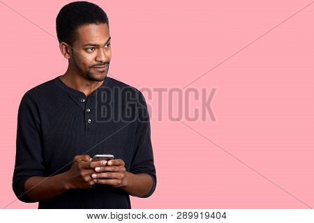 Image Of Handsome Guy With Dark Skin, In Black Shirt, Stands And Holds Phone In Hand, Looks Aside Wi