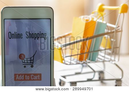 Smart Phone With Goods And Text Add To Cart On Screen And Paper Shopping Bags In Trolley Behind. Con