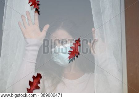 Woman In Medical Mask Indoors Looking Through Glass Window Looks At Falling Autumn Leaves. Autumn Co