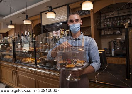 Cheerful Male Baker Wearing Medical Face Mask, Working At Hic Coffee Shop Selling Delicious Desserts