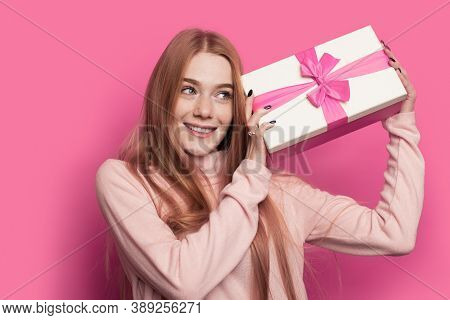Lovely Ginger Woman With Freckles And Red Hair Is Shaking A Present And Smile On A Pink Studio Wall