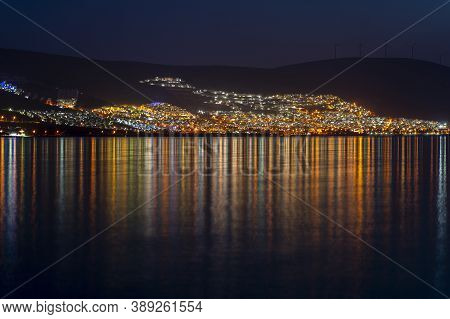 Night View Of The Turkish City Didim On The Aegean. Reflection Of Glowing Lights In Sea Water.
