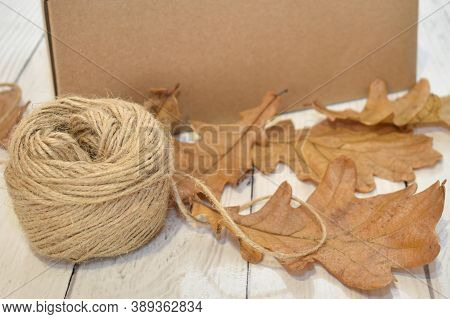 Cardboard Box, Autumn Oak Leaves And A Ball Of Rope On The Background Of A Wooden Table Top. Concept