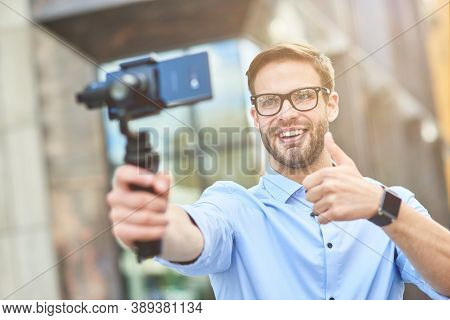 Young Cheerful Male Blogger Wearing Blue Shirt And Eyeglasses Holding A Gimbal With Smartphone, Smil