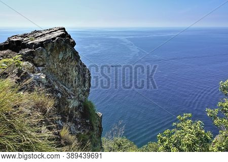 A Flat-topped Rock Juts Out Over The Calm Turquoise Sea. Green Plants Are Visible Below. Summer Sunn
