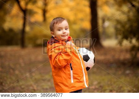 Little Football Player: Toddler Boy In Orange Jacket Standing Half Turned And Holding Soccer Ball In