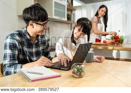 Asian little girl helping dad to work with computer laptop while mom cooking in kitchen. Family Work at home while quarantine from COVID-19. Happy family togetherness concept.