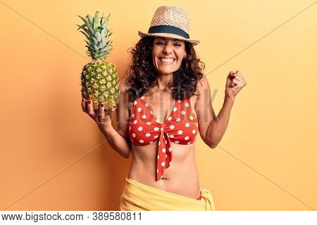 Middle age beautiful woman wearing bikini and summer hat holding pineappel screaming proud, celebrating victory and success very excited with raised arm 