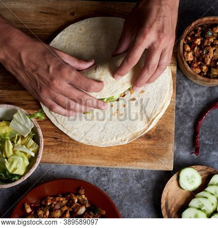 high angle view of a young caucasian man preparing a durum or a burrito, with chicken meat cooked with different vegetables such as onion or red and green pepper, and fresh lettuce and raw cucumber