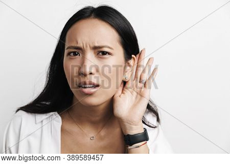 Displeased asian woman overhearing while posing at camera isolated over white background
