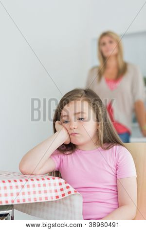 สาวน้อยนั่ง exasperated ที่แม่อำนวยตารางห้องครัวในพื้นหลังขณะ