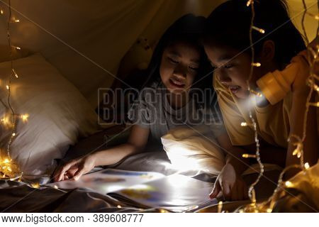 Family Concept. Elder Sister And Sister Reading Book With Flashlight Together Before Bedtime. Sister