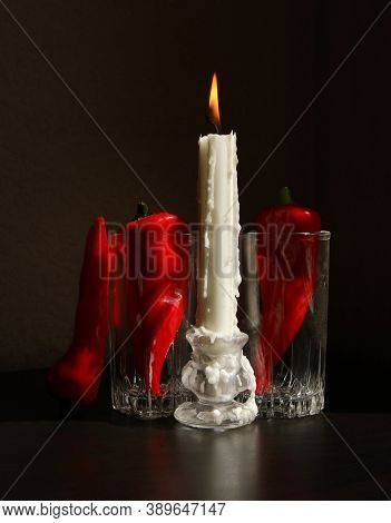 Still Life With Burning Candle In The Candlestick And Red Ramiro Peppers In The Glass Beakers Agains