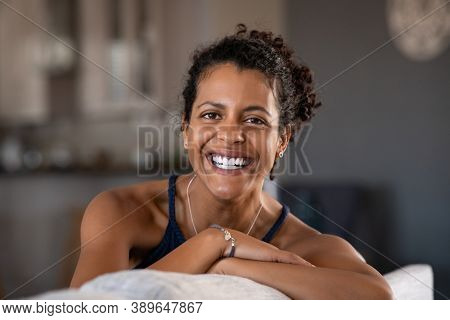 Cheerful ethnic mid adult woman relaxing on sofa at home. Successful young african woman with toothy smile sitting on couch and looking at camera. Portrait of confident black woman laughing.