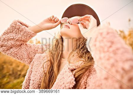 Autumn fashion. Stylish young woman in a fashionable fur coat and hat looks at the sky through pink glasses in an autumn park.