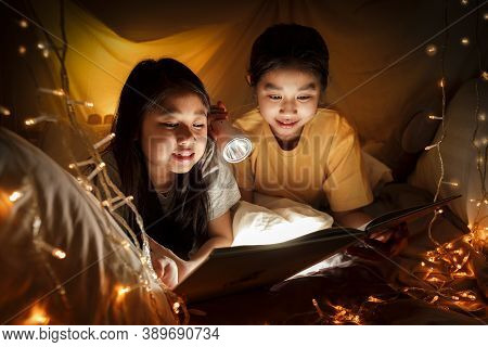 Family Concept. Elder Sister And Sister Reading Book With Flashlight Together Before Bedtime. Sister