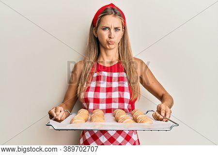 Young blonde woman wearing baker uniform holding homemade bread puffing cheeks with funny face. mouth inflated with air, catching air. 