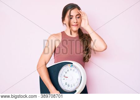 Beautiful young caucasian woman holding weight machine to balance weight loss stressed and frustrated with hand on head, surprised and angry face 