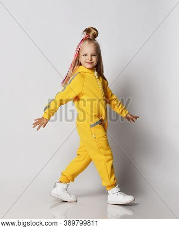 Playful Smiling Blonde Kid Girl With Girlish Hairstyle And Colorful Braids In Yellow Jumpsuit With H