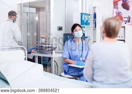Disabled Elderly Man In Wheelchair On Hospital Corridor Talking With Dorctor And Wearing Face Mask A
