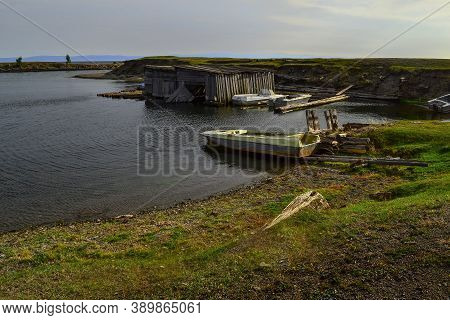 Old Small Rusty Iron Boats On Green Grassy Shore Of Bay, With Wood Barn On The Water. Lake Baikal, B