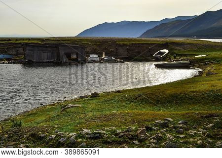 Old Small Rusty Iron Boats On Green Grassy Shore Of Round Bay, With Barn On The Water. Blue Baikal M