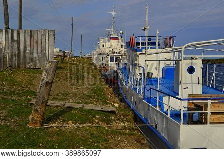 Large White Blue Boats, Ship  Tied To Pole In Green Grassy Shore With Wood Rustic Fence In Light Of 