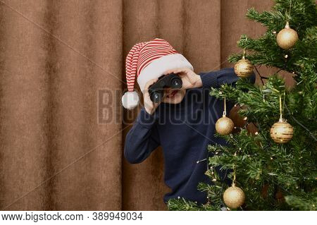 A Boy Peering Out From Behind A Decorated Christmas Tree, And Looks Through Binoculars. On A Brown C