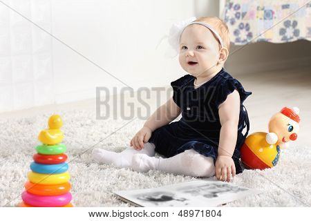 Little Cute Baby In Dress Sits On Soft Carpet Among Toys And Smiles.