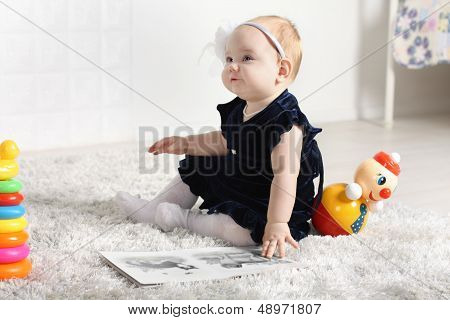 Little Cute Baby In Dress Sits On Soft Carpet Among Toys And Looks Up.