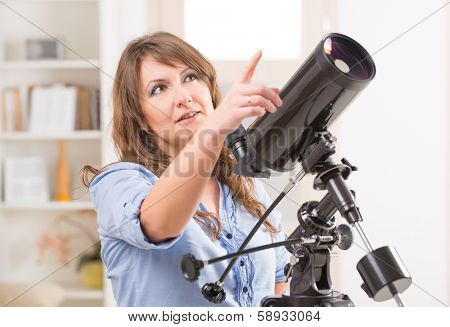 Beautiful woman looking skyward and pointing something in the sky standing near a window with astronomical telescope