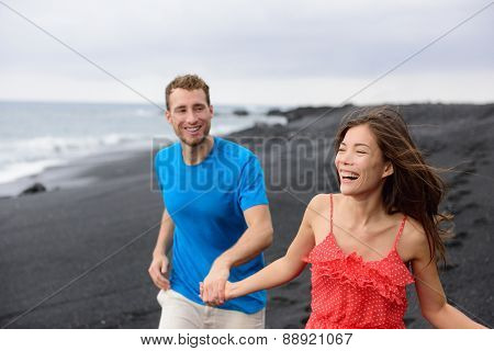 Happy couple laughing together walking on beach holidays. Vacation concept of two adult people holding hands on secluded black sand beach on volcanic island enjoying their getaway holiday.