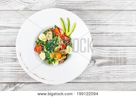Fresh salad with tuna spinach arugula codfish artichokes quail eggs avocado cherry tomatoes physalis and sesame on wooden background close up. Mediterranean food