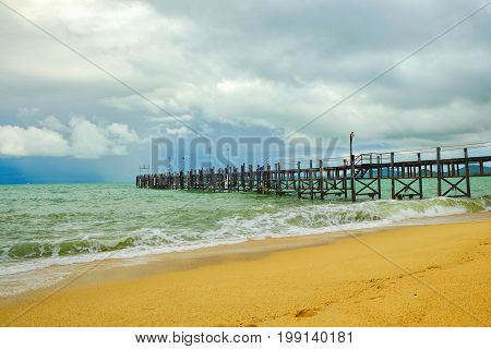 Old wooden pier stretching out to sea and foamy waves on the Maenam Beach, Koh Samui, Thailand.