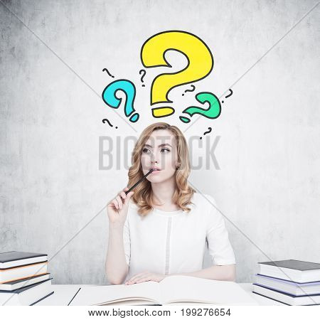 Beautiful young woman is sitting at a table with piles of book on it. The one in front of her is open. She is thinking and biting a pencil. Concrete wall with blue and yellow question marks