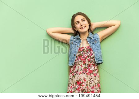 The beautiful long-haired blonde girl in flower dress having fun crossed hands onder head front of the light blue wall. Isolated studio shot.