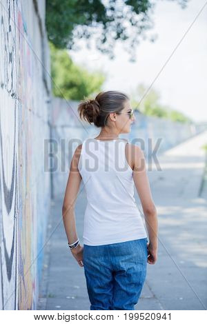 Back view of Hipster girl wearing blank white t-shirt, jeans and sunglasses posing against rough street wall, minimalist urban clothing style, mockup for tshirt print store