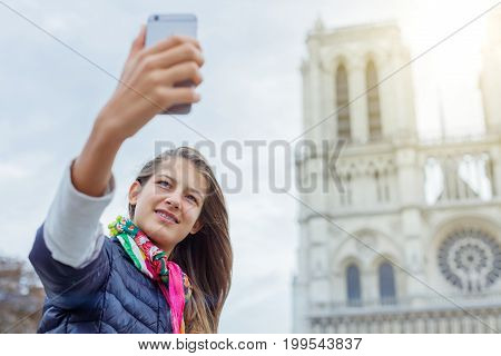 Teenager tourist girl is taking selfie on cathedral of Notre Dame de Paris with mobile smart phone. Travel, tourism, vacation concept. France.