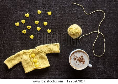 Cup of coffee, yellow knitted sweater for a doll and a heart-shaped buttons on a grunge black background.